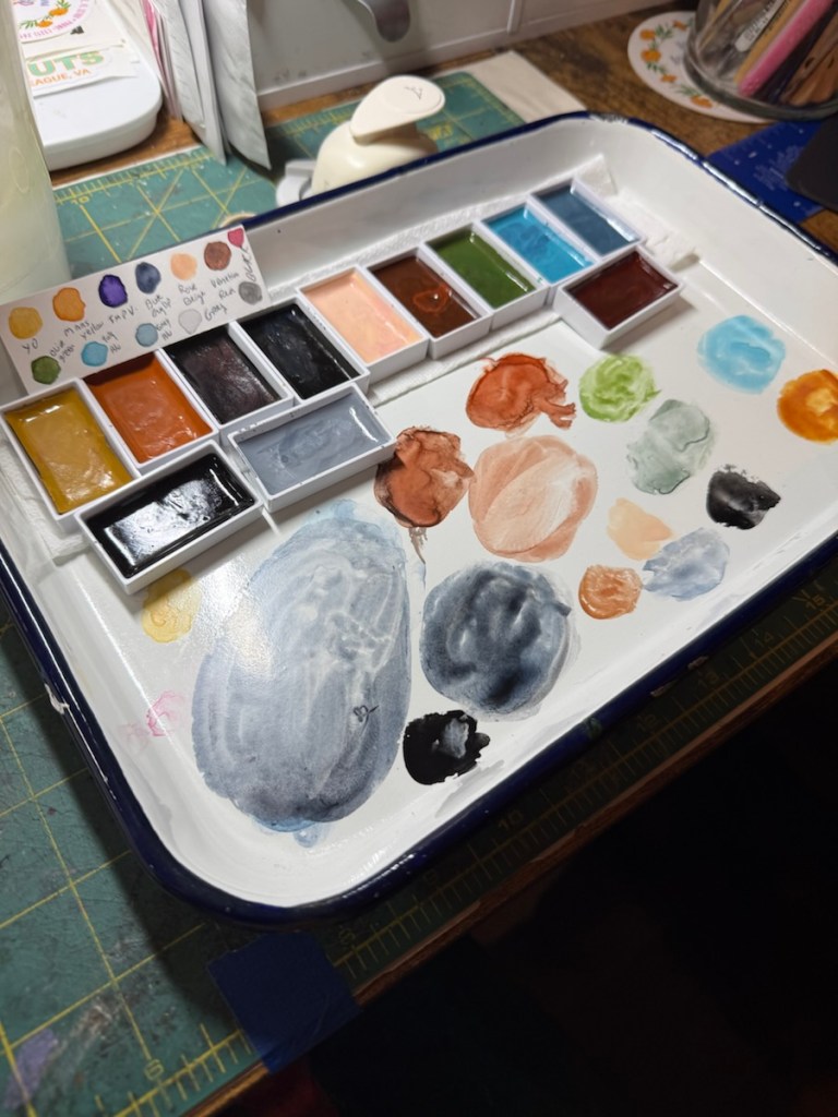 A watercolor palette featuring multiple wells filled with various paint colors, some partially mixed on a white tray, with a color chart at the top showing different shades.